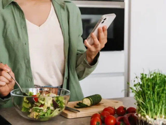 Mãos segurando o celular sobre a bancada, com salada e legumes sendo preparados